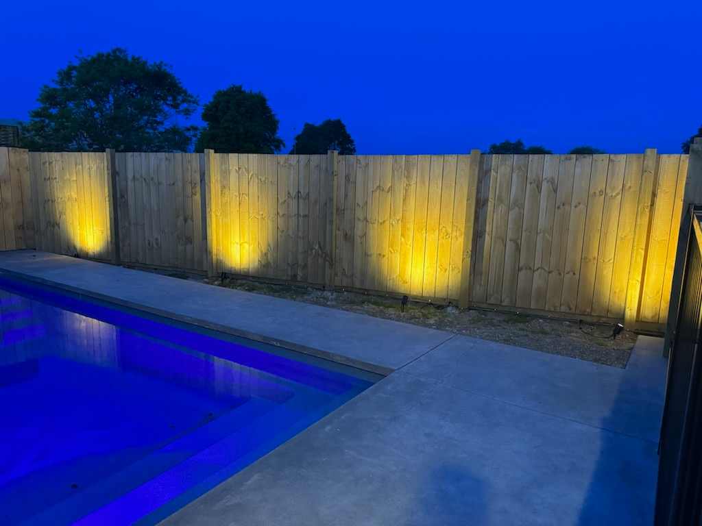 Garden spike lights washing a timber fence beside a swimming pool at dusk